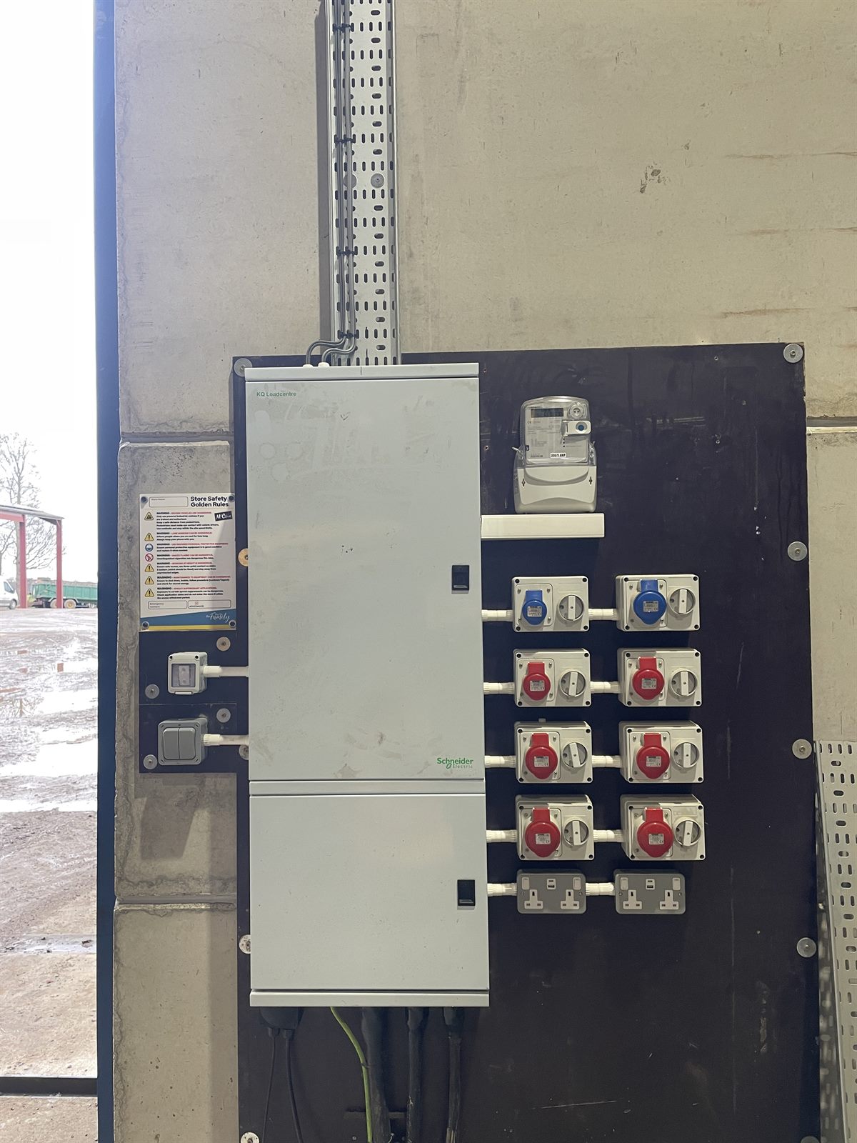 A three phase distribution board in a farm building: Schneider KQ Loadcentre on a backboard with a three phase meter above, six red three phase sockets, two blue 16A single phase sockets and two twin 13A sockets, each with its own isolator.