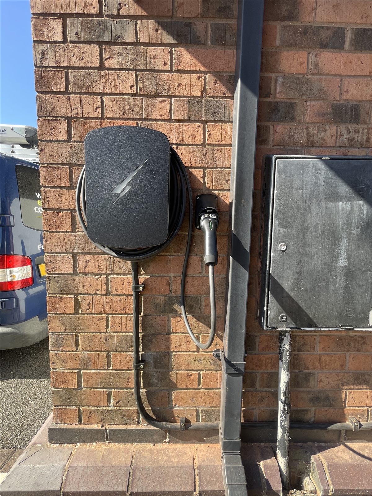 Close view of a wall-mounted home EV charger with the charging cable coiled and the connector holstered, cable clipped neatly down the brickwork.